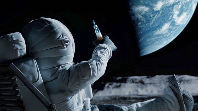 An astronaut holds up a drink to the Earth while sitting in a chair