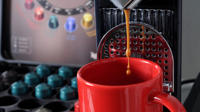 Nespresso machine extracting coffee from Nespresso pod into red coffee mug beside case of Nespresso pods