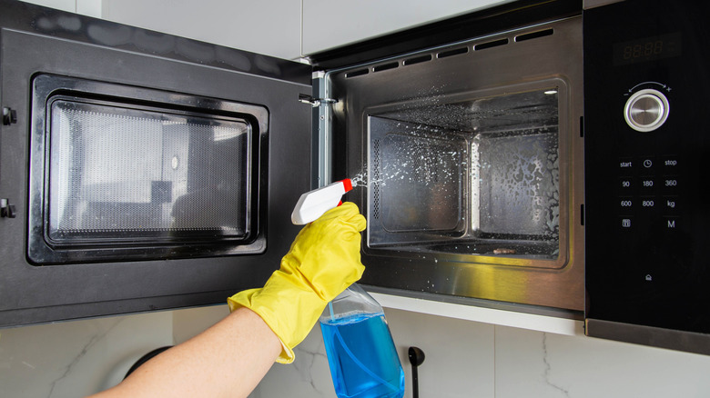 A hand in a yellow rubber glove holding a bottle of spray and cleaning the interior of a microwave