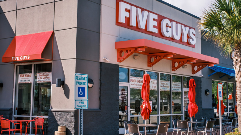 The exterior of a Five Guys restaurant with tables and chairs with red umbrellas outside