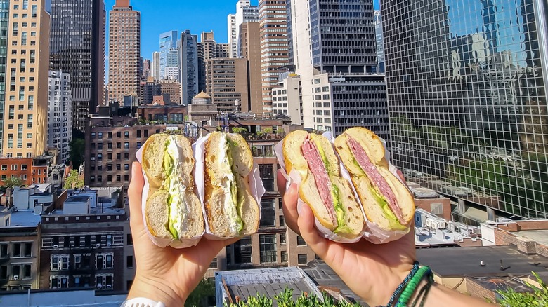 Hands holding bagel sandwiches with city backdrop