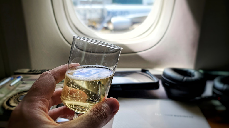 person holding glass of white wine in first class flight beside window in airplane