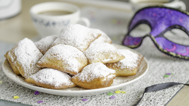 beignets on plate with a cup of coffee and a mardi gras mask in the background