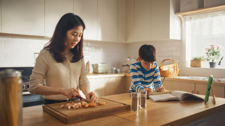 An Asian woman using a wooden cutting board in the kitchen witrh her son nearby doing homework