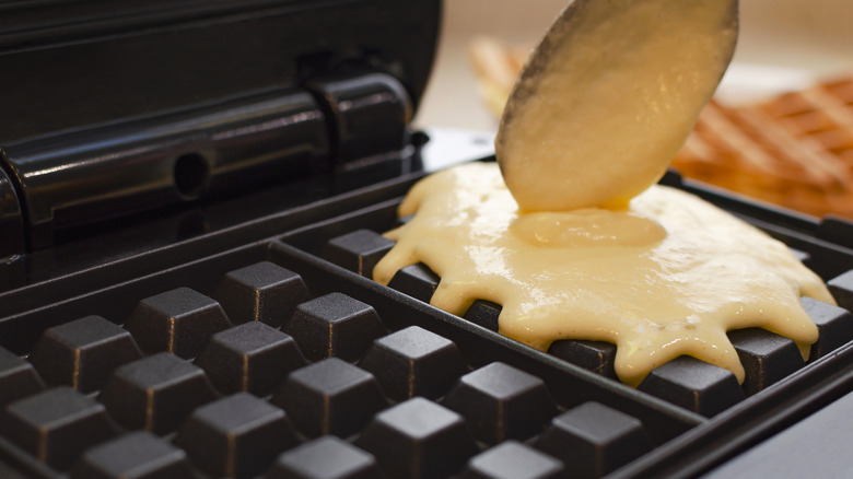 A person ladling batter into a waffle maker with a wooden spoon