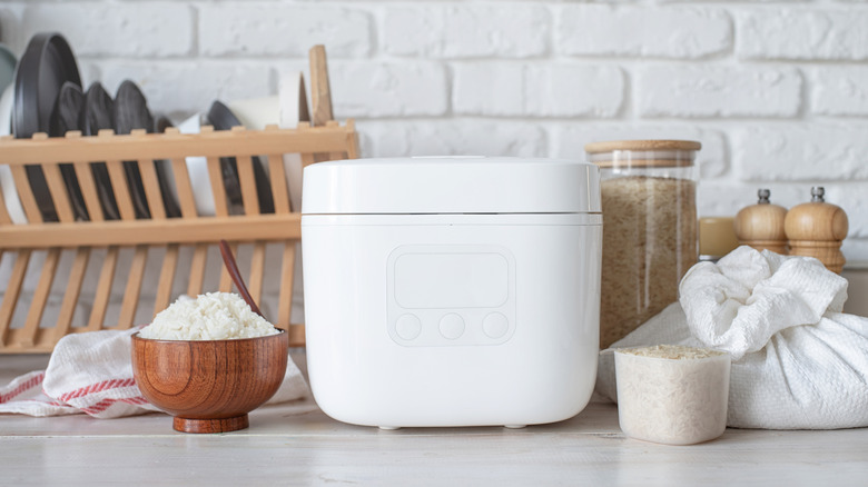 A white rice cooker with bowl of cooked and uncooked rice sitting next to it.