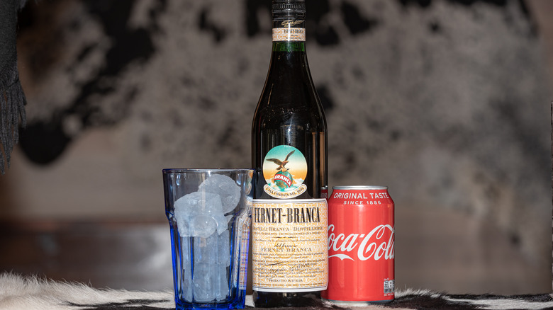 A bottle of Fernet-Branca with a Coke can and a glass of ice cubes