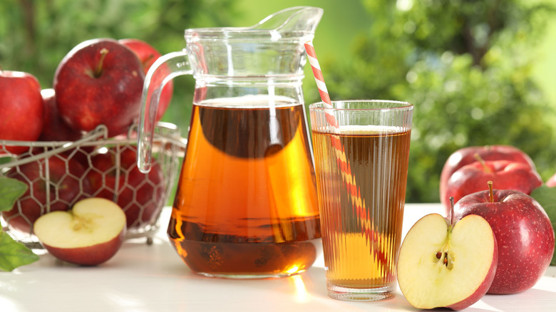 A pitcher of apple juice beside a glass of apple juice and a basket of apples