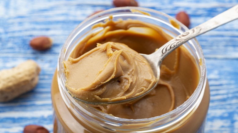 A jar of peanut butter scooped with a spoon, with peanuts in the background