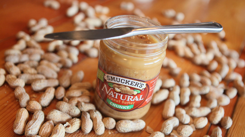 A jar of Smucker's Natural Peanut Butter with a knife, surrounded by peanut shells