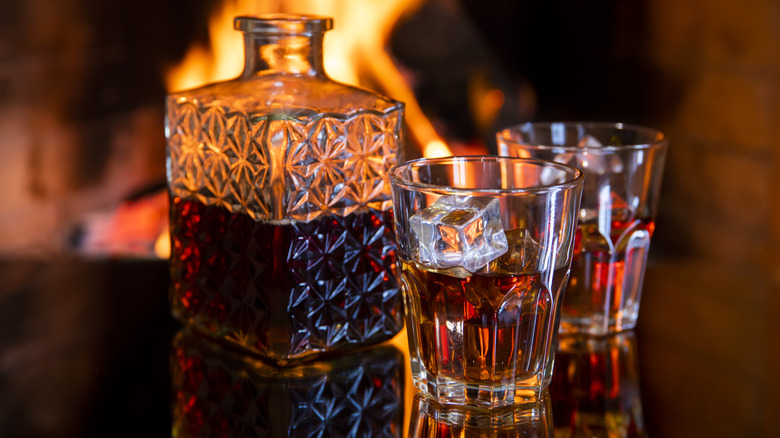 A glass decanter of whiskey with two glasses in front of a fire