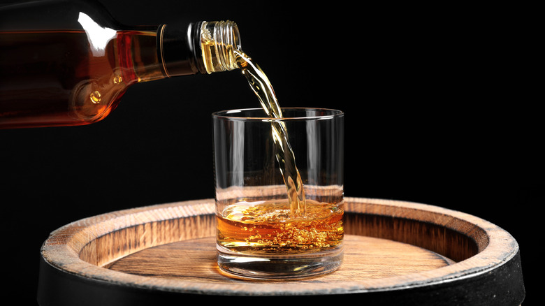 Whiskey being poured from a bottle into a glass on a wooden barrel