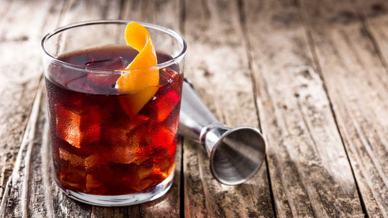 A boulevardier with an orange garnish rests near a jigger cup on a wooden table