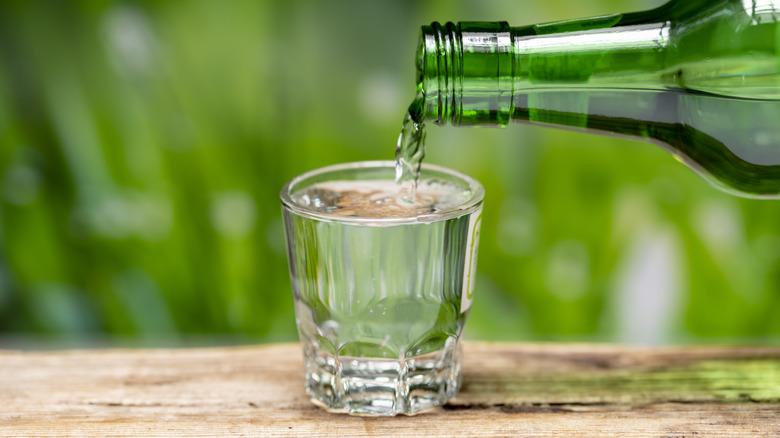 Soju from a green bottle being poured in a clear shot glass on a wooden surface