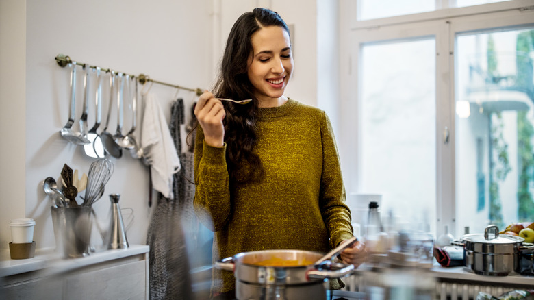 a dark-haired woman tasting soup from a pot on the stove