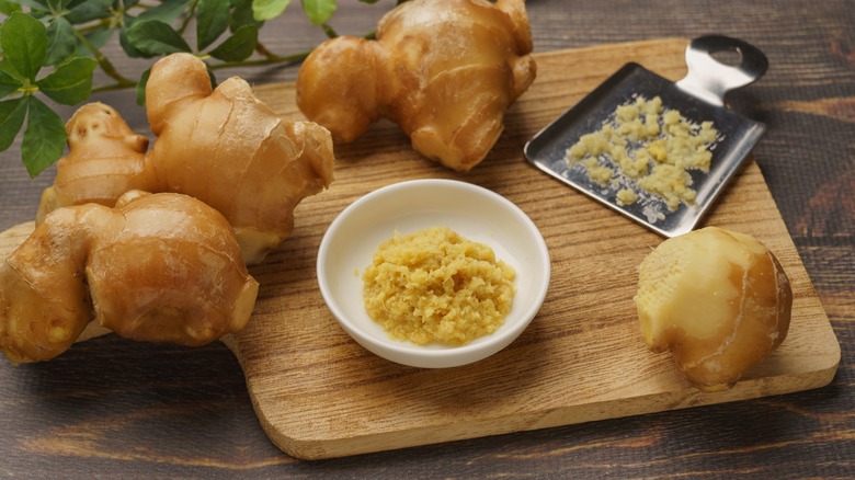 A small bowl of grated ginger, with whole ginger root and a gingery cutting board in the background.