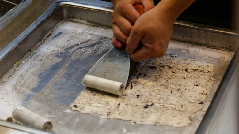 A person rolling cookies and cream ice cream off a frozen sheet pan with a metal spatula.