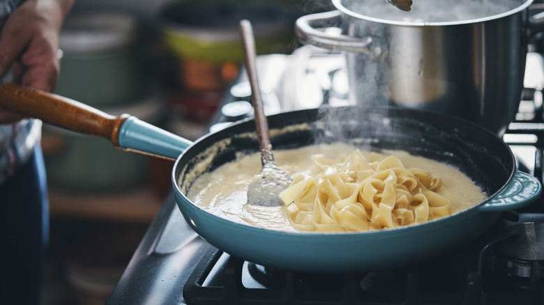 Stirring large fettuccine noodles into a large frying pan filled with Alfredo sauce