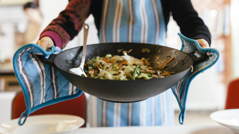 A person holding a large wok filled with a dish that resembles stir fry