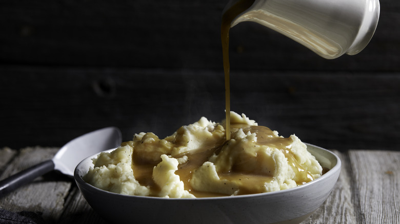 Pouring brown gravy over a large bowl of hot mashed potatoes.