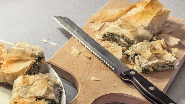 Serrated bread knife with a spinach roll on a wooden cutting board.