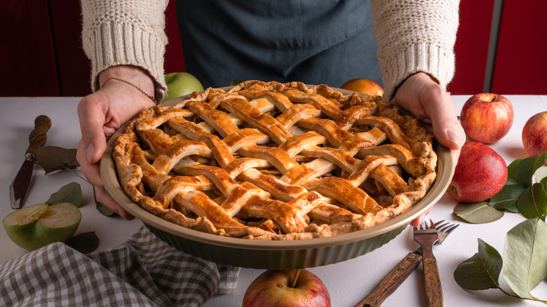 A person holding an apple pie in a dish with latticed pastry on top