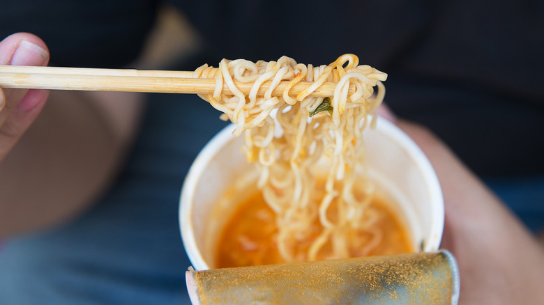 Instant ramen is in a styrofoam cup as someone holds up ramen noodles on chopsticks