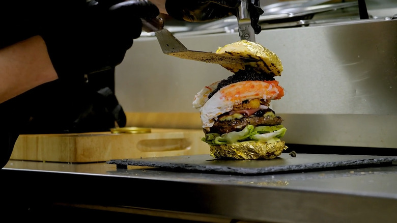 Chef putting on a gold-flaked top bun onto a burger sitting on a counter