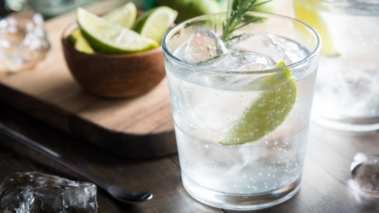 A rocks glass with vodka and soda, with bowl of limes in background.