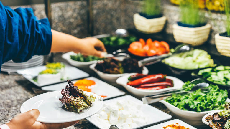 Person grabbing some food in a buffet line with a plate of lettuce in the other hand