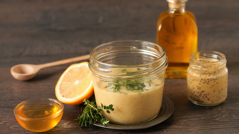 A jar of homemade honey mustard next to fresh thyme, honey, a lemon slice, Dijon mustard, and olive oil