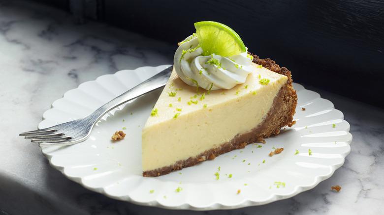 Slice of key lime pie on a small white plate with a lime garnish and whipped cream on top