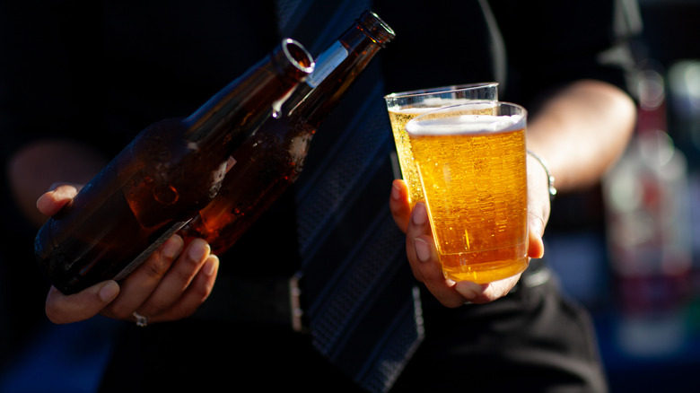 A person holding two empty bottles of beer in one hand and two cups full of beer in the other