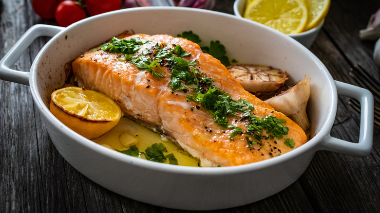 Cooked salmon in a white pan alongside lemons, garlic, oil, and herbs.