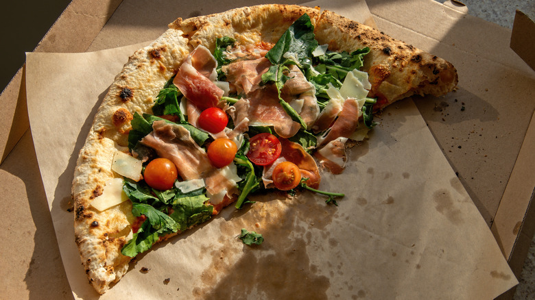 Half a pizza with tomatoes, cheese, prosciutto, and spinach in an open pizza box