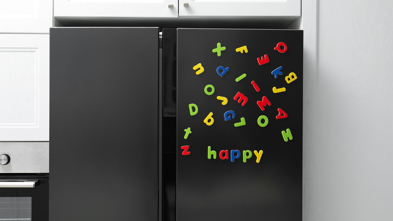 A black fridge with colorful letter magnets on the door in a kitchen with white cabinets.