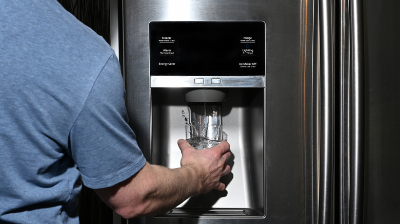 Male right hand filling glass with water splashing out of dispenser of home fridge