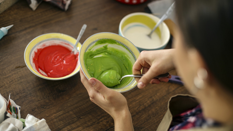 A person mixing green food dye in a small bowl with a dish of red food dye nearby