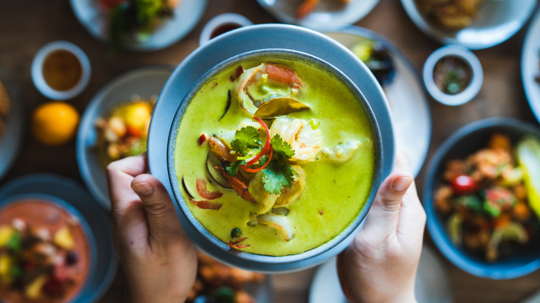 hands holding a bowl of Thai green curry with chicken and vegetables above table of various traditional Thai dishes