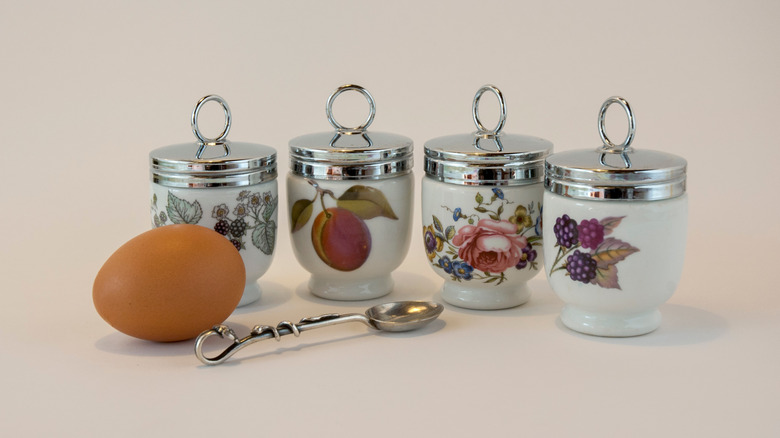 Four porcelain coddlers with metal lids and a small spoon and whole egg nearby