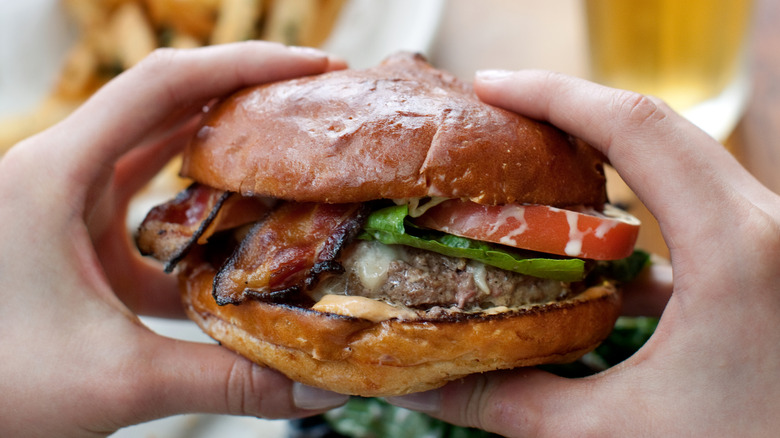 A person holding a hamburger in a bun with bacon, lettuce, and tomato