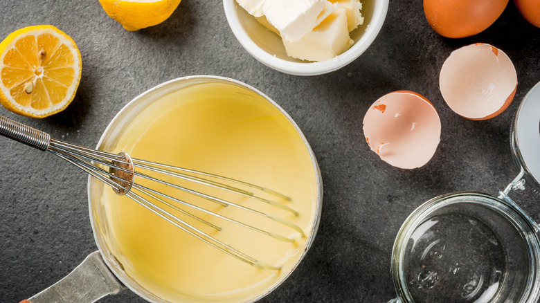 A bowl of hollandaise sauce with a whisk sits on a black surface surrounded by lemon, butter, and eggshells