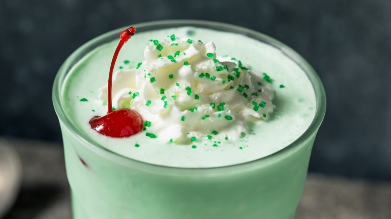 Green milkshake in a tall glass with whipped cream and a cherry on top, along with two spiral straws and a four leaf clover decoration all on a dark, texturized backdrop.