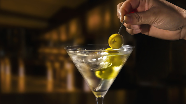 A hand holding an olive to a martini glass