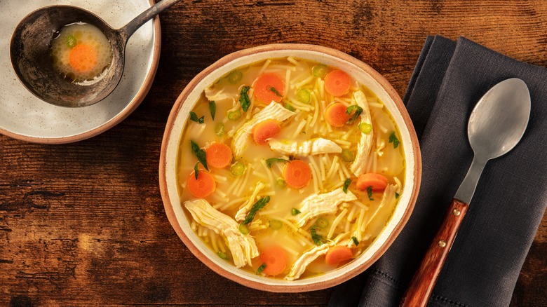 Bowl of chicken noodle soup on a wooden table with ladle and spoon