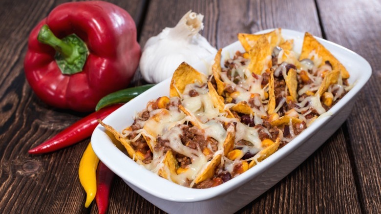 Nachos covered in melted cheese in a baking dish, next to peppers and garlic on a wooden table