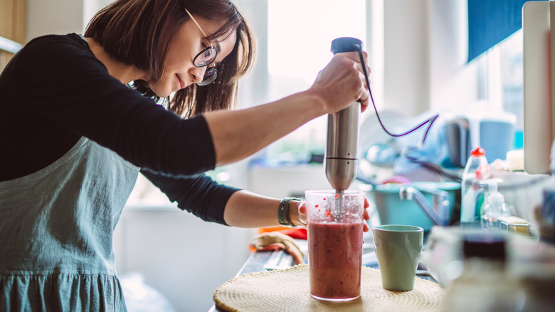 A woman with glasses using a hand mixer to make a fruit smoothie in a cup