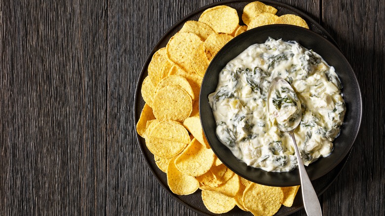 A bowl of artichoke dip with potato chips on the side