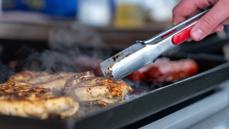 A hand holding tongs grilling chicken on griddle