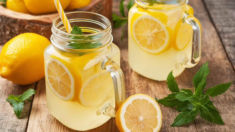 two mason jars filled with freshly squeezed lemonade with herb garnish surrounded by lemons on wooden table